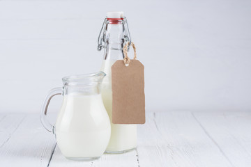 Bottle of milk and jar of milk with blank cardboard label on white wooden table background