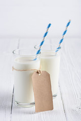 Two glasses of milk with blue stripped paper straw and blank paper tag on rope on white wooden table still life