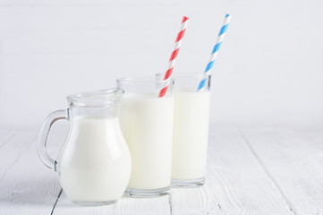 Glasses of milk with stripped blue and red paper straws and jug of milk on white wooden table still life