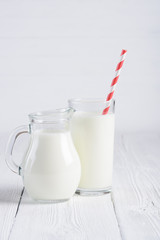 Glass of milk with stripped red paper straw and jug of milk on white wooden table