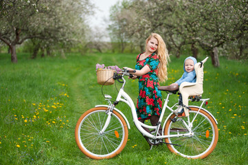 Fototapeta premium Smiling mother in flowered dress holding bike with child who sits in bicycle chair, against the background of blooming trees, dandelions and fresh greenery in spring garden