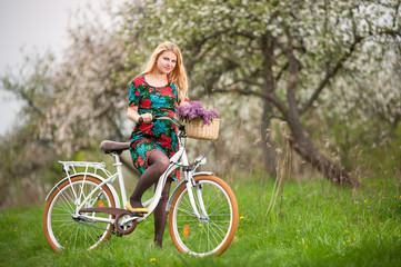 Young woman with long blond hair wearing flowered dress and yellow shoes starting to ride a vintage white bicycle with flowers basket