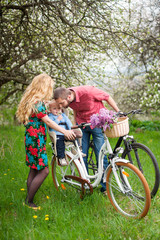 Fototapeta na wymiar Young family on a bicycles in the spring garden. Mother holding your bike and baby sitting in bicycle chair, in the basket lay a bouquet of lilacs, against the background of blooming fresh greenery