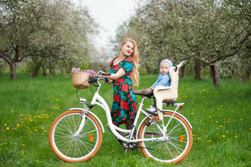 Fototapeta premium Happy mother in flowered dress holding bike and looking at child who sits in bicycle chair, against the background of blooming trees, dandelions and fresh greenery in spring garden