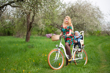 Mother with long blonde hair in dress keeps bicycle and baby in bicycle chair, in the basket lay a bouquet of lilacs, against of blooming fresh greenery in spring garden on the background