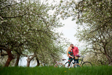 Fototapeta premium Young couple of bikers loving and keeping bikes against the background of blooming trees and fresh greenery in spring garden. Couple together enjoying romantic holidays. Side view
