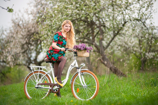 Young female biker with long blond hair wearing flowered dress and yellow shoes riding a vintage white bicycle with flowers basket, against the background of blooming trees in spring garden