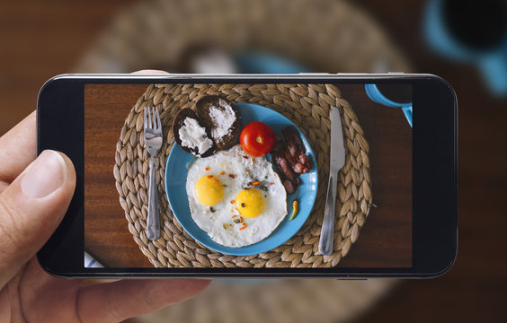 Taking picture of fried eggs and tomato with mobile phone. Phone in male hands.On the plate there is 2 fried eggs, tomato, bread with cheese. fork and knife. Vintage style.