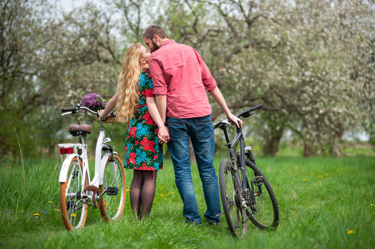 Young couple in love standing with the bicycles back to camera, holding hands and kissng each other in the spring garden, against the background of trees and fresh greenery.