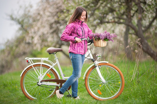 Beautiful brunette girl wearing purple jacket and jeans with a vintage white bicycle and lilac flowers basket, against the background of blooming trees, fresh greenery in spring garden