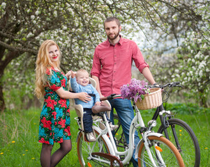 Obraz premium Beautiful family on a bicycles in the spring garden. Mother holding her bike and baby sitting in bicycle chair, in the basket lay a bouquet of lilacs, against the background of blooming trees