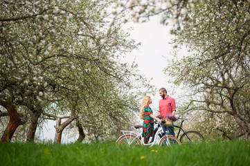 Loving happy couple with bikes looking at each other against the background of blooming trees and fresh greenery in spring garden. Couple together enjoying romantic holidays. Side view