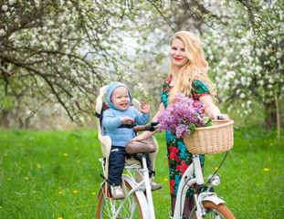 Beautiful young mother with long blonde hair and red lips holding vintage bike and happy baby sitting in bicycle chair against the background of blooming fresh greenery in spring garden
