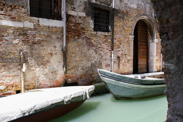 Fototapeta na wymiar Two boats in the narrow channel of water a milky green. Old brick wall with crumbling plaster, arched doorway.