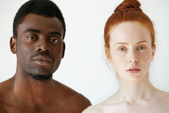 Love against racism. Black and white. Coffee and milk. Headshot of young loving multi-ethnic couple standing shirtless against white studio wall, looking at the camera. African man and Caucasian woman