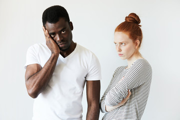 Portrait of young Caucasian female with red hair, standing with arms folded, angry and disappointed with her African boyfriend who looking at the camera with tired expression fed up with quarrelling