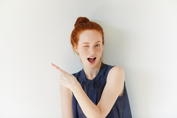 Portrait of young redhead female with perfect freckled skin showing something interesting at copy space wall for your information or advertising content, winking at the camera and pointing sideways