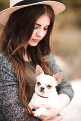 Smiling young girl wearing hat holding chihuahua outdoors. Woman with pet dog.