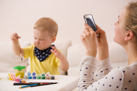 Young mother taking photo of cute little toddler boy in yellow shirt sitting at table and drawing with colorful paints. Early learning. Creative. Toddler drawing. Mom making picture on mobile phone