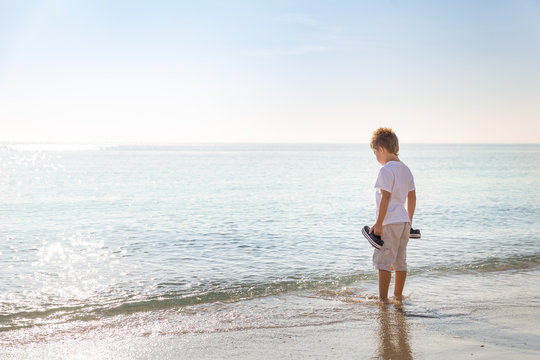 Adorable Kid Boy Standing On The Sandy Beach By The Ocean And Looking On The Gorgeous Summer View. Miami Summer Vacations. Blue Sky With Clouds As A Background. Child On Amazing Beach.