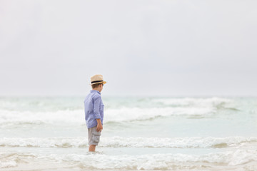 Portrait of adorable kid boy in straw hat and sun glasses walking on ocean beach and looking at the waves. Vacations by the sea. Outdoor.