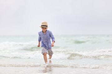 Portrait of adorable kid boy in straw hat and sun glasses running on ocean beach and looking at the waves. Vacations by the sea. Outdoor.