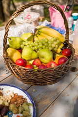 Basket with organic fruit (apples, pears, bananas and grapes) on the wooden table with nuts and other ingridients for putdoor picnic. Healthy food for summer picnic.