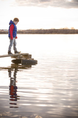 Cute kid boy standing on a wooden bridge and looking at the water in a cloudy spring or autumn day. Child on the river shore. Outdoors. on the pontoon bridge on the river