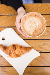 Top view on woman's hand holding big cup with cappuccino coffee and fresh French croissant on a wooden table in outdoor cafe. Breakfast with coffee and bread.