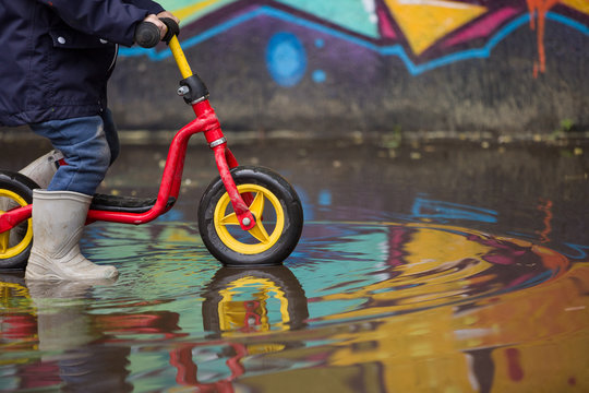 Little Child In Grey Rubber Boots Cycling After Rain.Toddler In A Pair Of Grey Rubber Boots In A Big Puddle With Grafiti Wall Reflection. Fun After Rain. Boy On The Bike On The Street.