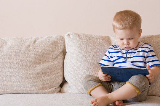 Adorable blond toddler boy laying on the sofa and playing with tablet pc at home, indoors. Child with tablet computer.