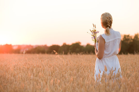 Back view on blond woman with flowers walking on the wheat field on sunset. Beautiful happy girl enjoying summer sun on the meadow. Lifestyle and summer concept. Outdoors.