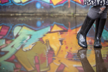 Girl in rubber boots standing in the puddle in the street. Woman in grey rubber boots splashing in a puddle after rain. Pair of grey rubber boots in a big puddle with graffiti reflections.