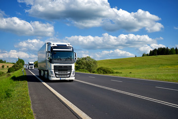 Three white trucks driving on asphalt road in a rural landscape. Mirroring the surrounding countryside on the container. Sunny day with blue skies and white clouds over green meadows.