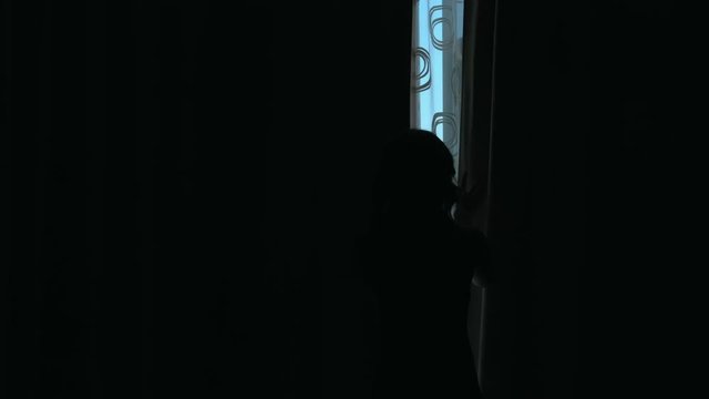 Silhouette of a girl who opens the curtains and looking out the window