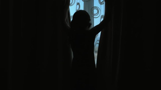 Silhouette of a girl who opens the curtains and looking out the window