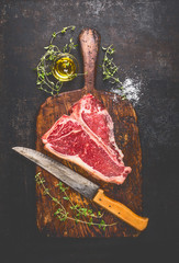 Raw T-bone Steak for grill or BBQ with fresh herbs and oil and kitchen knife on dark aged cutting...