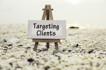 conceptual image with word TARGETING CLIENTS on white canvas frame with wooden tripod stand.Blurred Clamshell and cockles background.