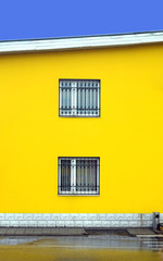 Part of yellow colored building wall with two windows closed metal grills vertical photo over blue cloudless sky