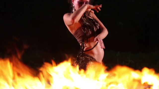 Attractive young woman dancing in the night with a fire flame in the foreground
