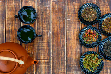 Different kinds of tea on plates near brown teapot and two cups with tea on wooden background. Assortment of dry tea. Tea concept. Tea leaves. Copy space