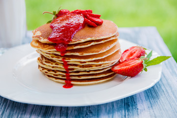 Pancakes with fresh strawberry and jem near glass with milk on white plate on white wooden background  in garden or on nature background.  Stack of pancakes on the table.