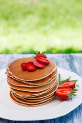Pancakes with fresh strawberry and mint on white plate on pink wooden background  in garden or on nature background.  Stack of pancakes on table.