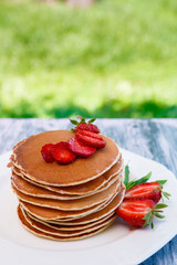 Pancakes with fresh strawberry and mint on white plate on pink wooden background  in garden or on nature background.  Stack of pancakes on table.