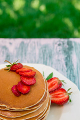 Pancakes with fresh strawberry and mint on white plate on pink wooden background  in garden or on nature background.  Stack of pancakes on table.