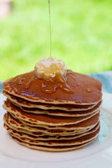 Pancakes with butter and honey on white plate on  in garden or on nature background. Maple syrup pouring on pancakes stack.