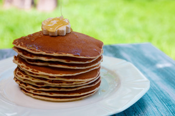 Pancakes with butter and honey on white plate on  in garden or on nature background. Maple syrup pouring on pancakes stack.