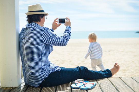 Rear side view of father with smartphone taking picture of baby. beach terrace on sunny day outside
