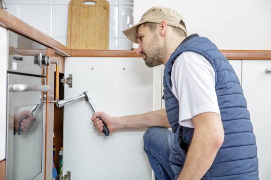 Worker repairing the sink in the kitchen. Man looks at the sink and holding a wrench.