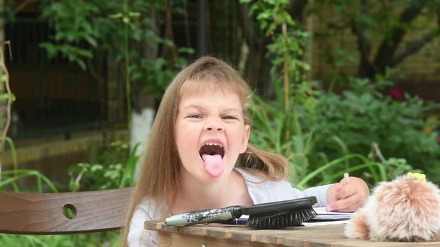 Girl sits at a table in the courtyard and grimacing showing tongue to the frame